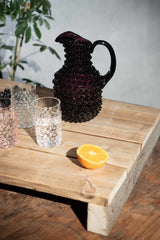 Violet Hobnail Square Jug on a wooden pallet surrounded by products from the Hobnai collection with a green plant and a concrete wall in the background
