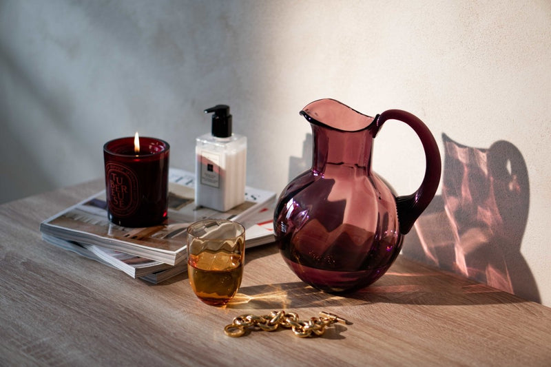 Violet Marika Jug on a wooden table surrounded by a candle, magazines, a gold jewel on a white background and shadows.